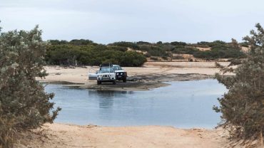 Off-road vehicles parked near water on a sandy riverbank, surrounded by native bushland, in a remote.
