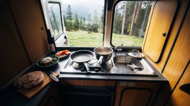 Compact campervan kitchen with stove and scenic forest view outside.