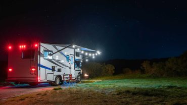 RV parked in open field at night with starry sky and trees in background.