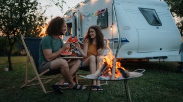 Family camping scene with children and a caravan, outdoor fire, and evening sky.
