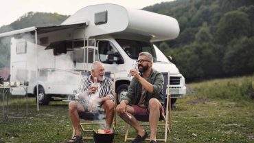 Camping with friends enjoying drinks near a motorhome in a lush green landscape.