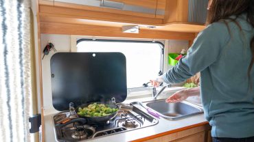 Kitchen in a campervan with stove, sink, and window view.