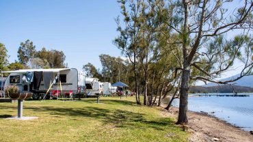 Camping by the water with caravans and trees in the background.