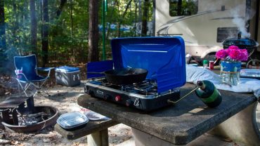 Camping stove with frying pan on outdoor table in forest setting.