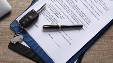 Close-up of a car key, pen, and signed documents on a wooden desk.