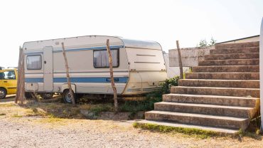 Vintage caravan parked near concrete stairs in outdoor setting, with wooden posts and gravel ground.