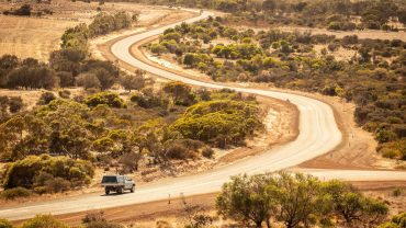 Winding dirt road in the Australian outback with sparse vegetation and a vehicle traveling along it.
